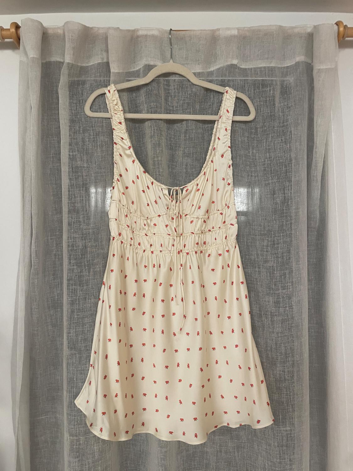 Cream dress with a small red floral print, hanging on a hanger in front of a gray translucent curtain.
