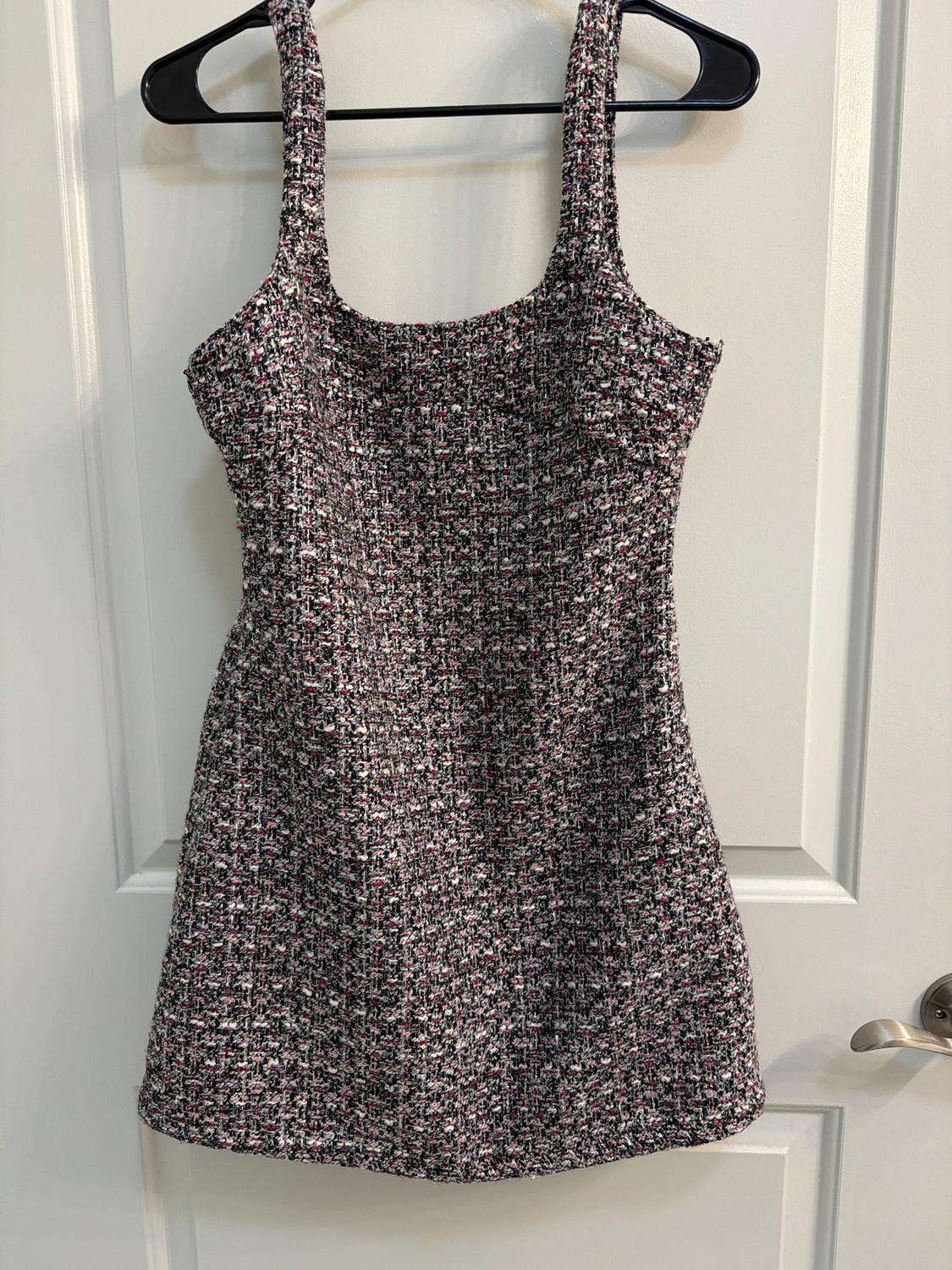 Sleeveless dress made of textured fabric in black, white, and pink tones, hanging on a black hanger over a white door.