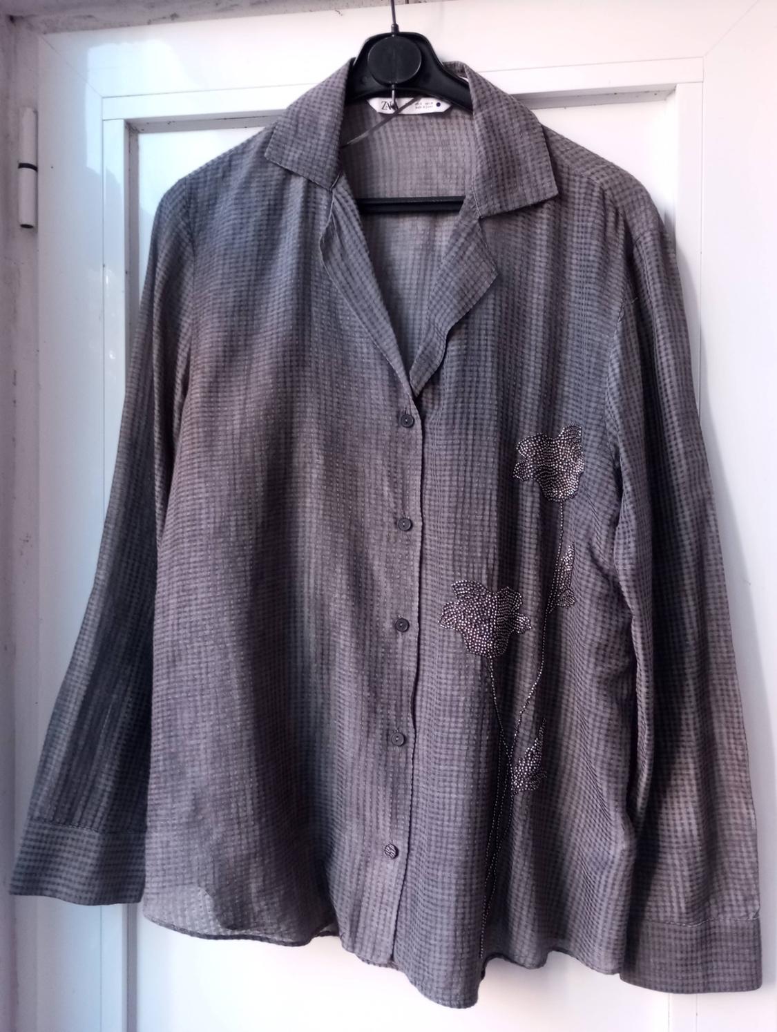 Small grey checked shirt hanging on a black hanger, with floral embroidery on the right side, on a white background.