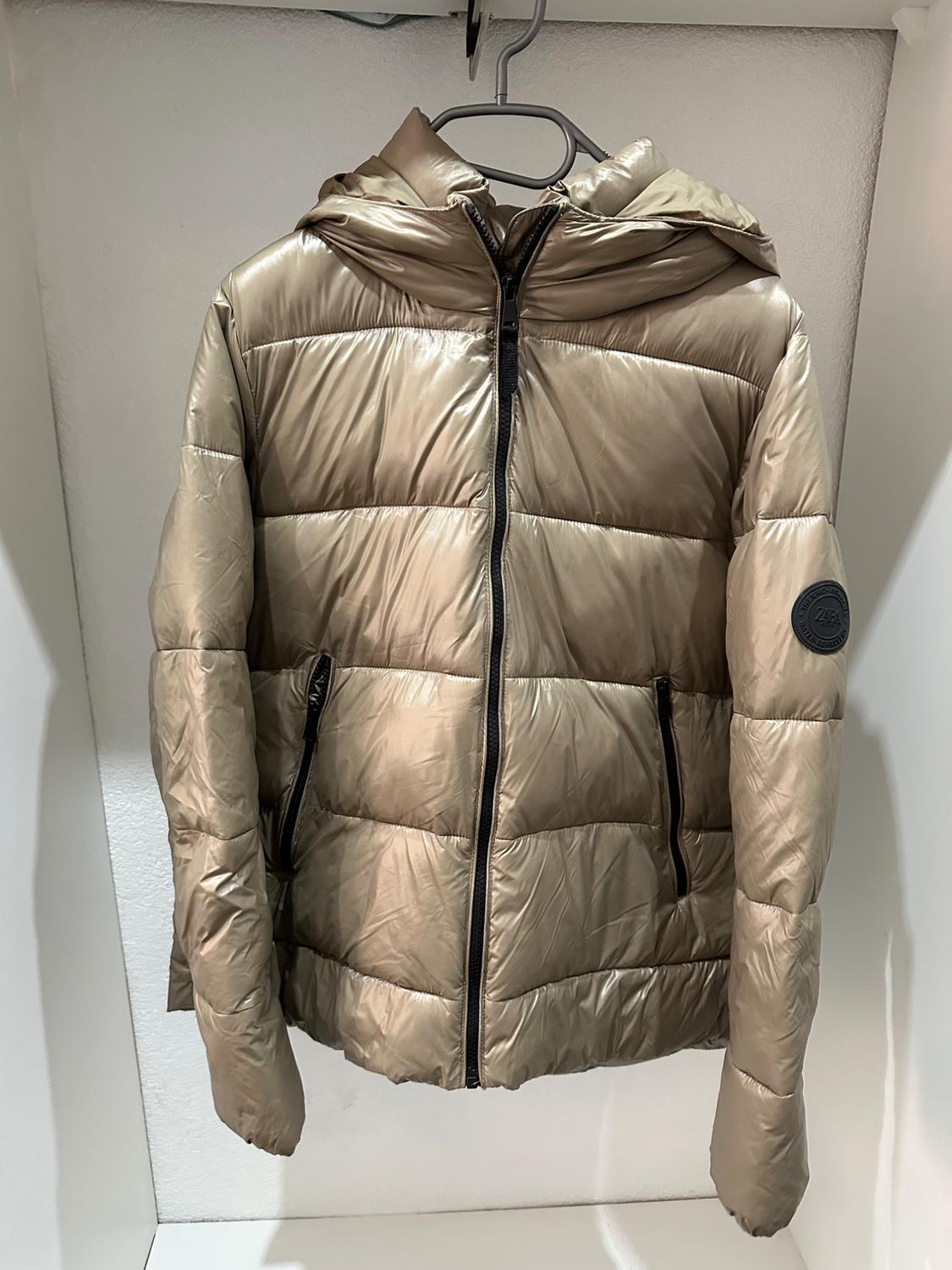 Beige padded jacket with a hood and black zip, hanging on a hanger inside a white wardrobe.