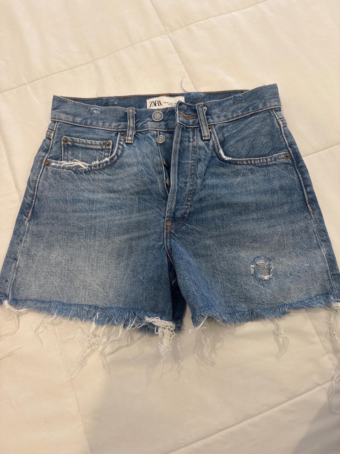 Blue denim shorts with frayed edges and a small tear on the right leg, on a white quilt.