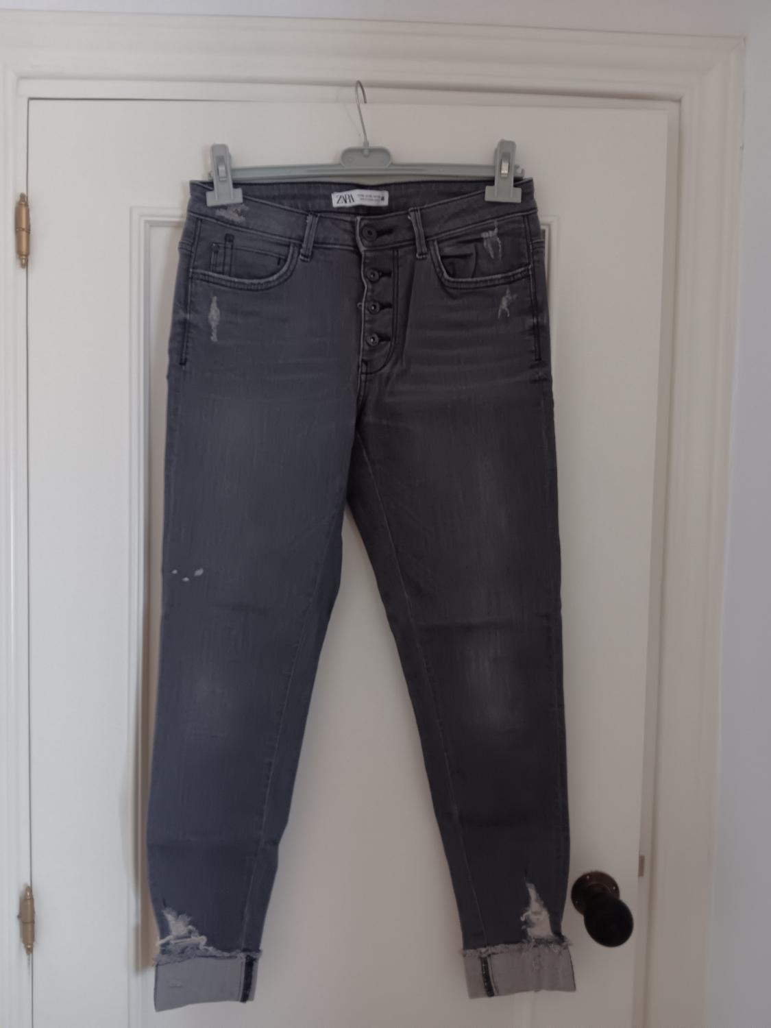 Worn grey jeans hanging on a white door, with visible buttons and frayed hem. "ZARA" label on the waistband.