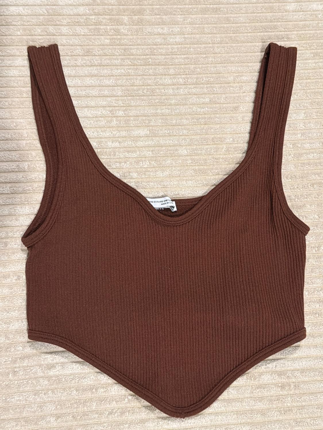 Brown ribbed cropped top with wide straps, a deep neckline, and a pointed hem, photographed against a textured beige fabric background.
