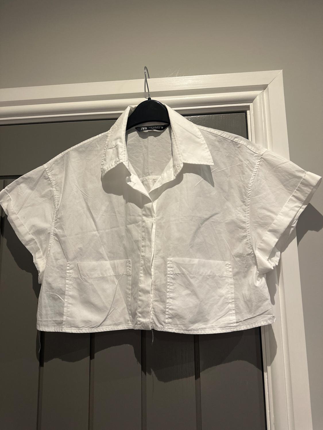 Short-sleeved white shirt hanging on a black coat hanger over a grey door. The shirt has two front pockets.