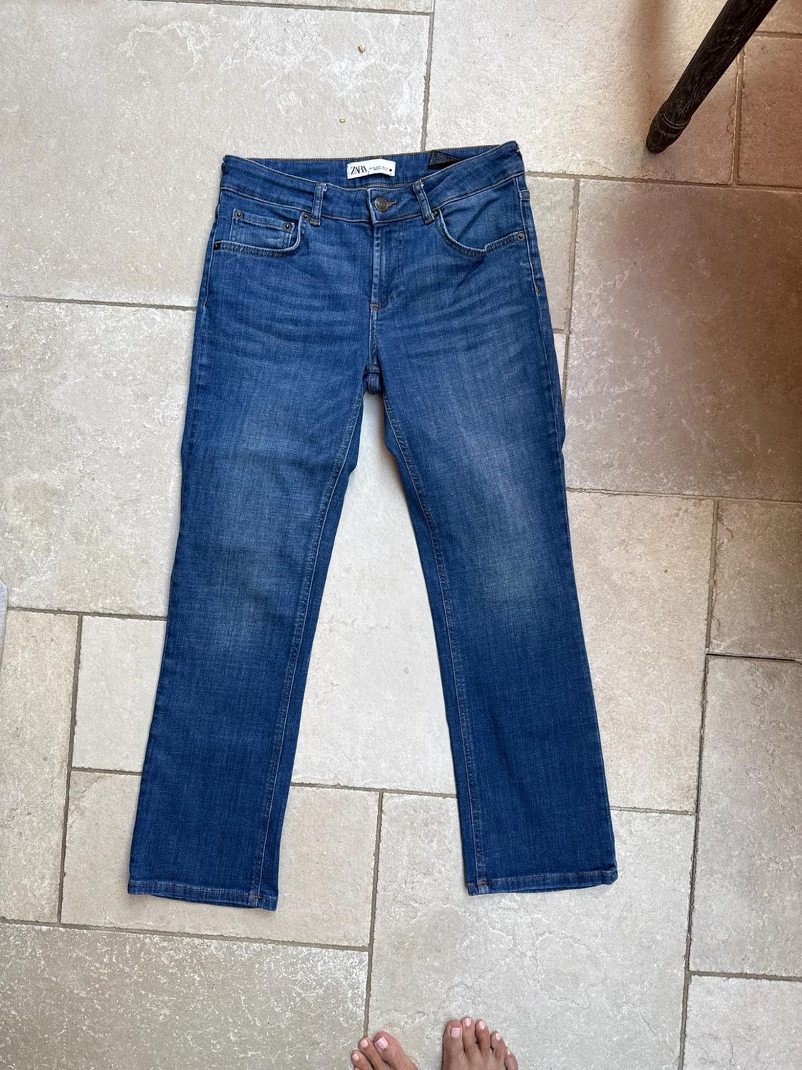Blue jeans with a ZARA label on a light-tiled floor. Bare feet are visible at the bottom edge.