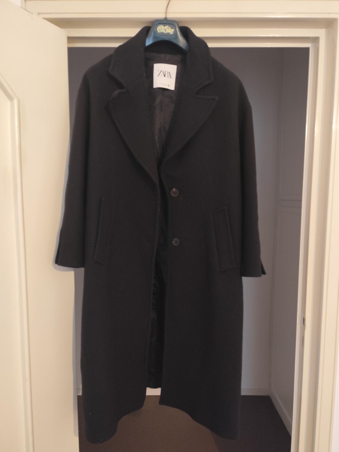 Black coat hanging on a door, with a visible label that reads "ZARA". The coat has wide lapels and two front buttons.