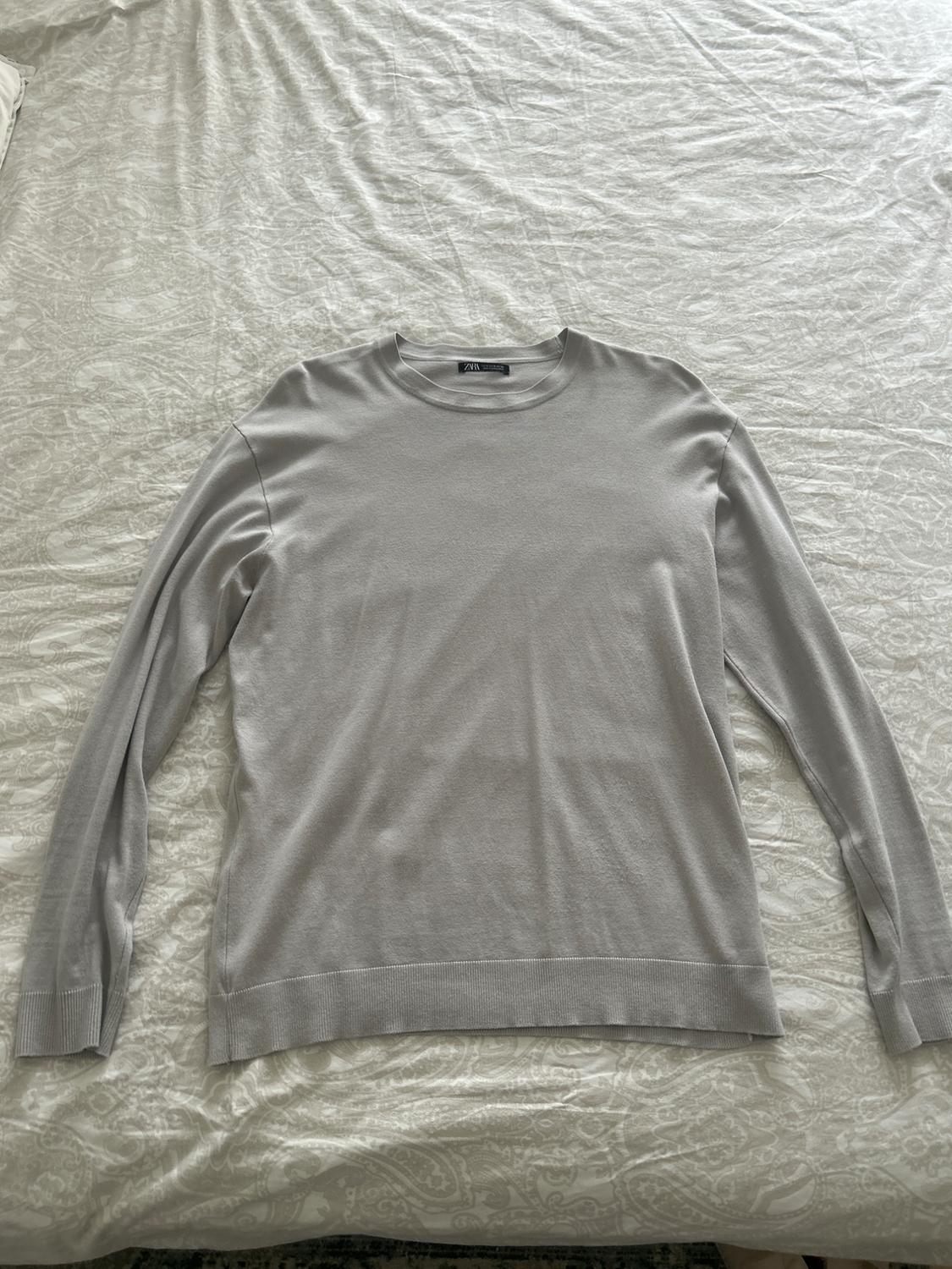 Light grey long-sleeved crew neck jumper on a bed with patterned sheets. The label reads ZARA.