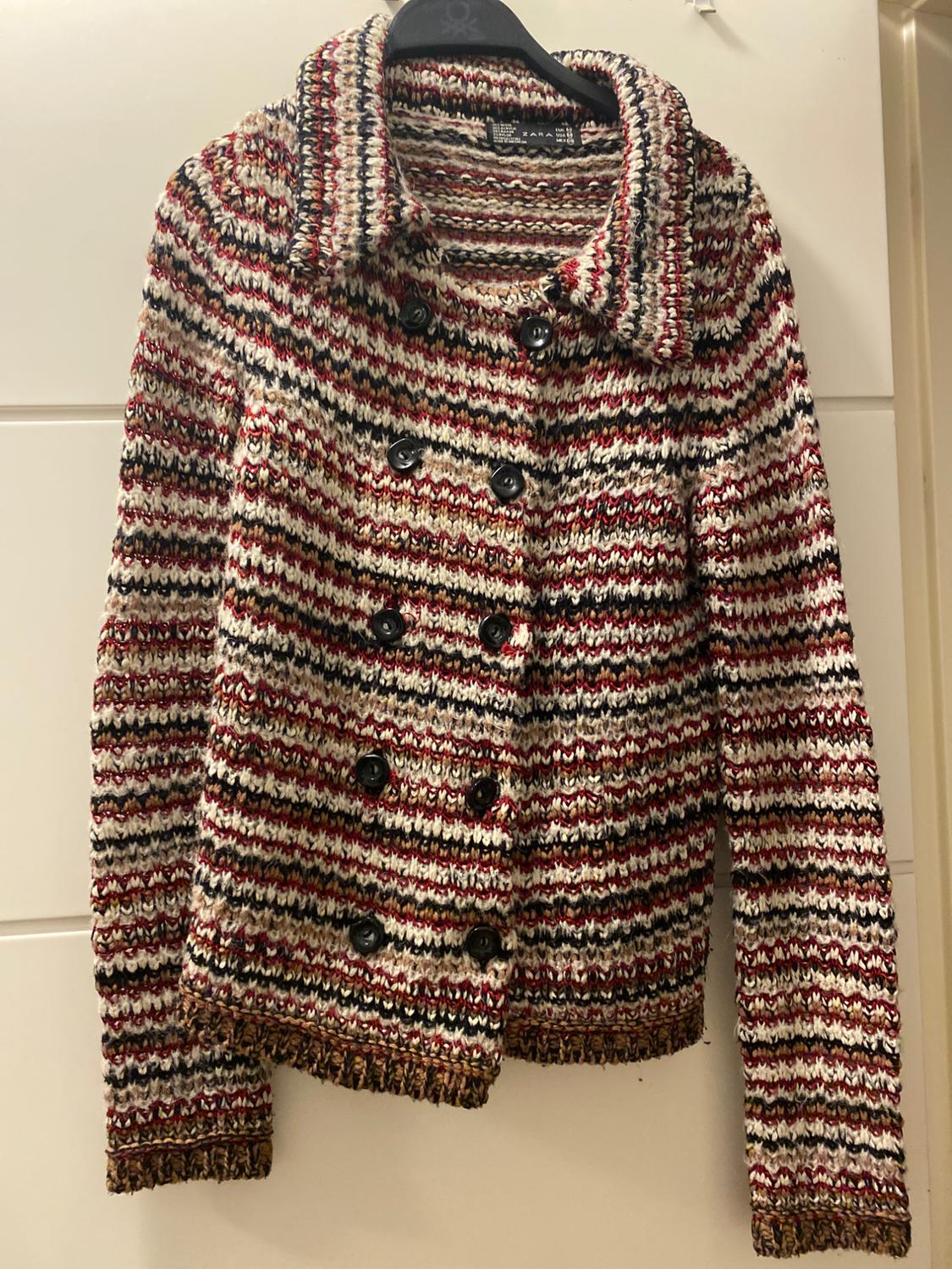 Multicoloured knitted jacket with horizontal stripes and black buttons, hanging on a black hanger against a white background.