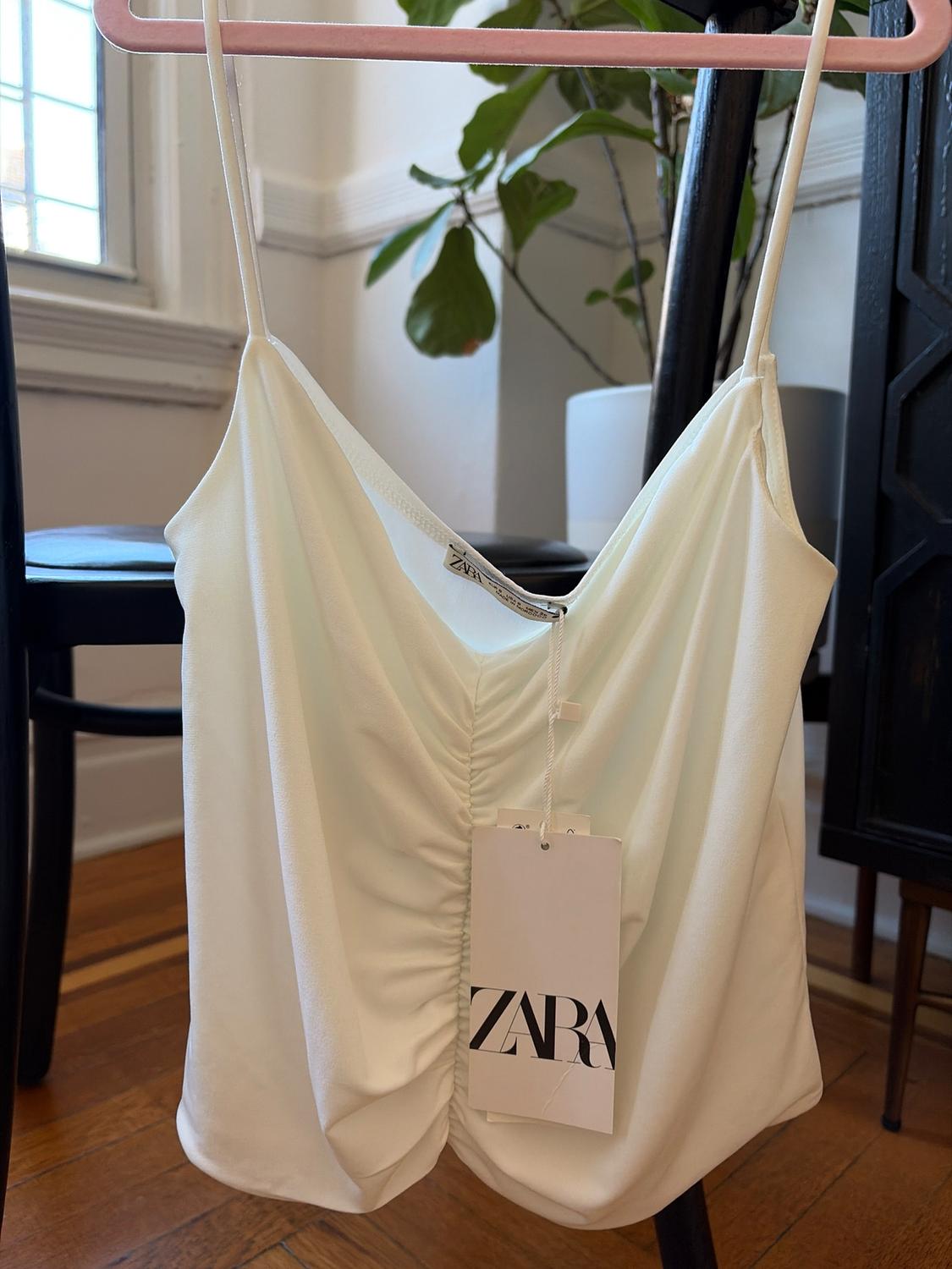 White top with thin straps and central ruching, hanging on a pink hanger. Visible label with the text "ZARA". Background with plant and window.