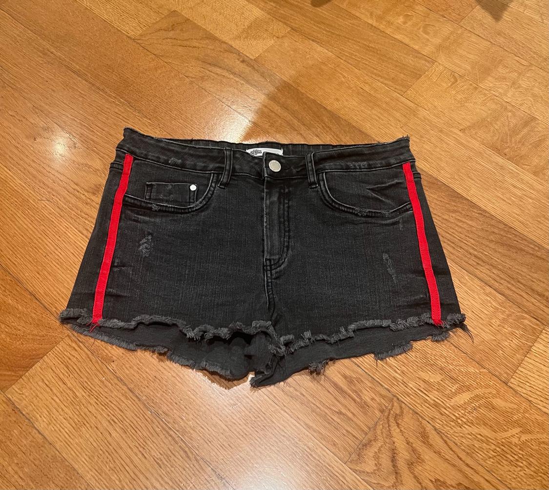 Black denim shorts with red side stripes and a frayed hem on a wooden floor.