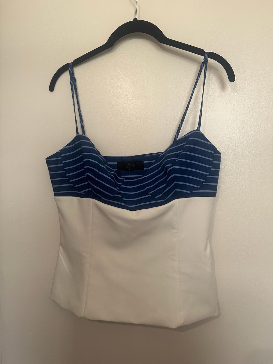 Cami top hanging on a black hanger against a white wall. Blue chest with horizontal stripes and a plain white body.