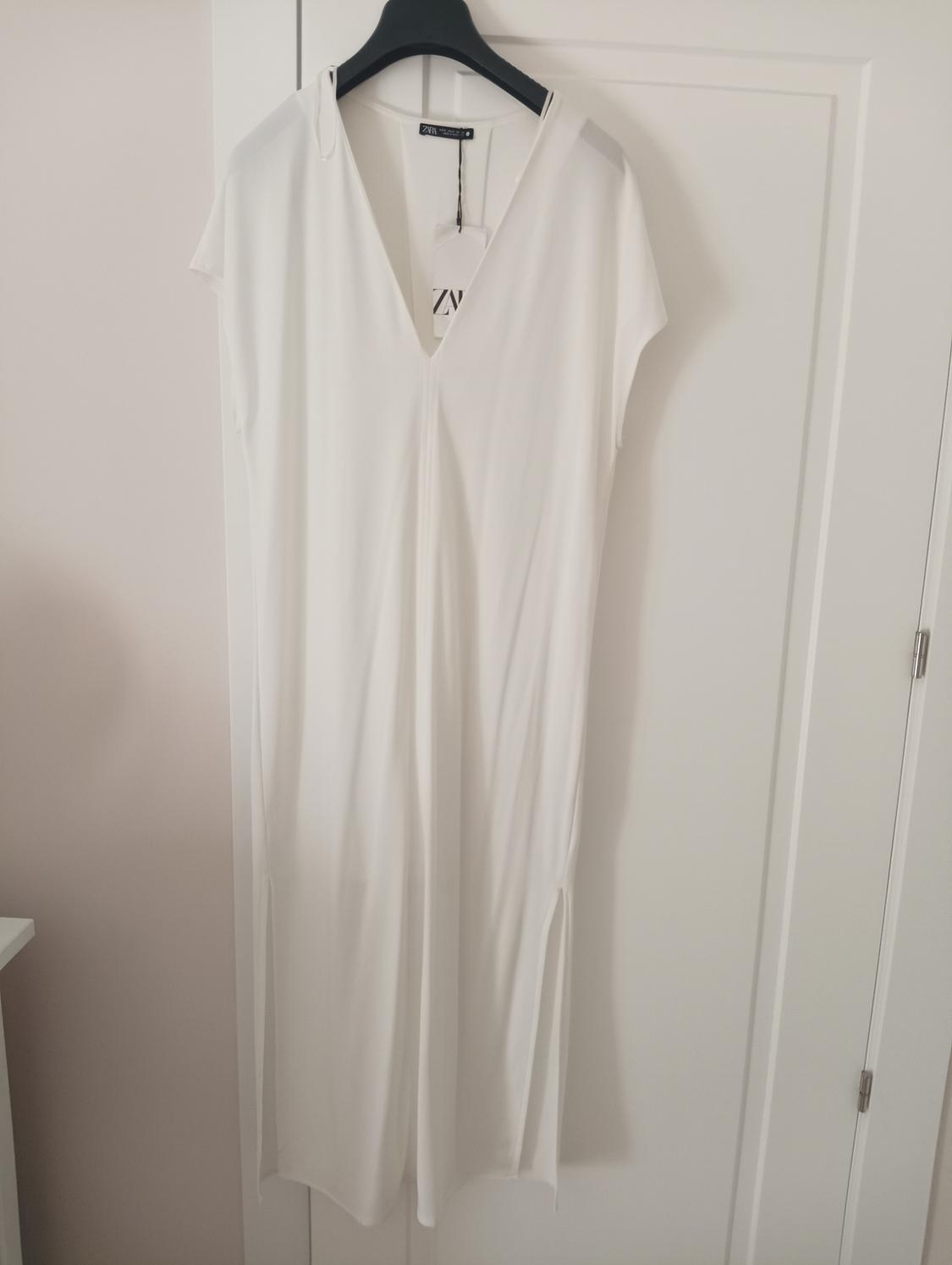 White short-sleeved dress hanging on a black hanger over a white door. Visible label with text "ZARA".
