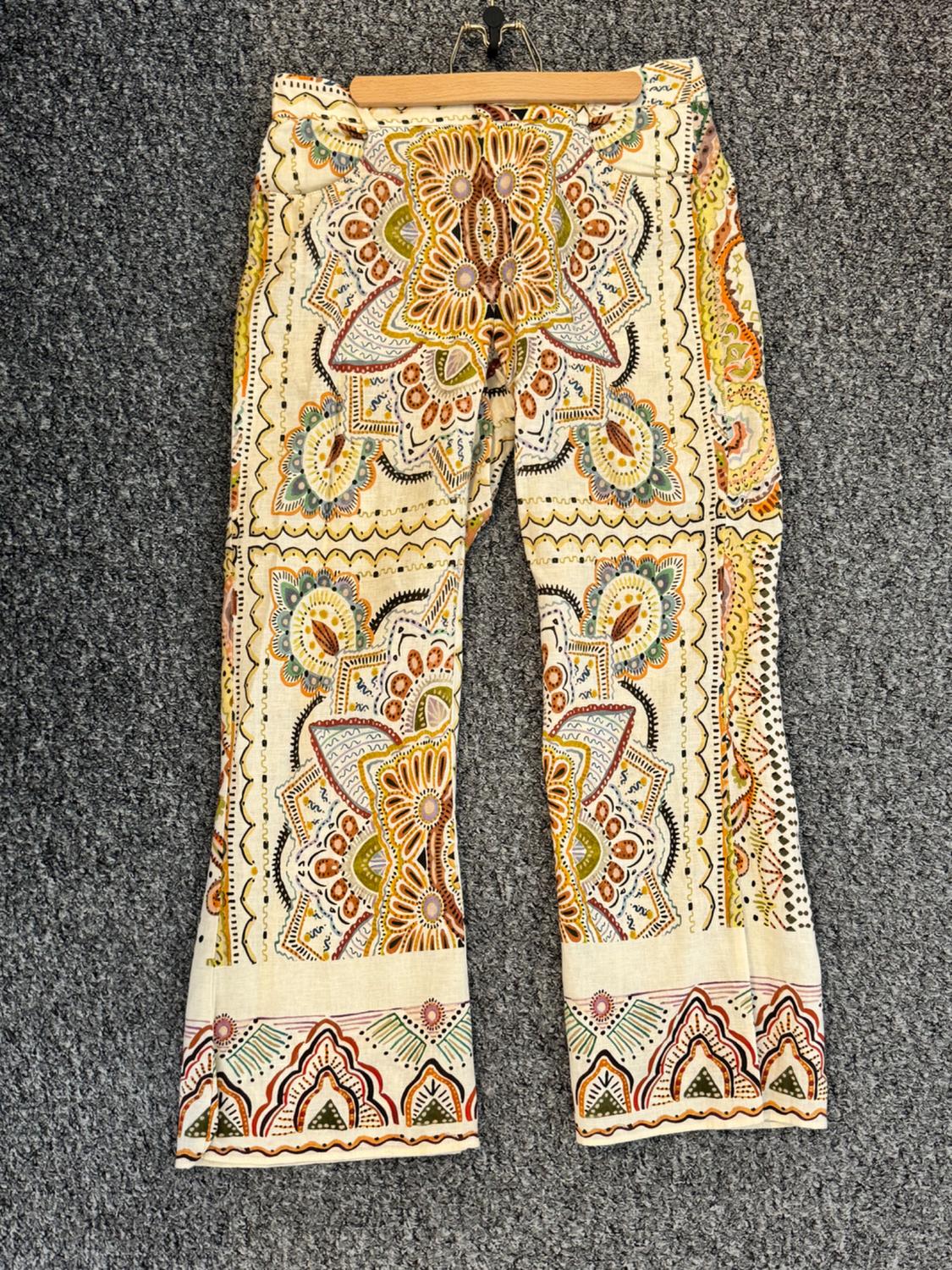 Beige trousers with floral and geometric print in shades of orange, green and yellow, hanging on a hanger against a grey background.