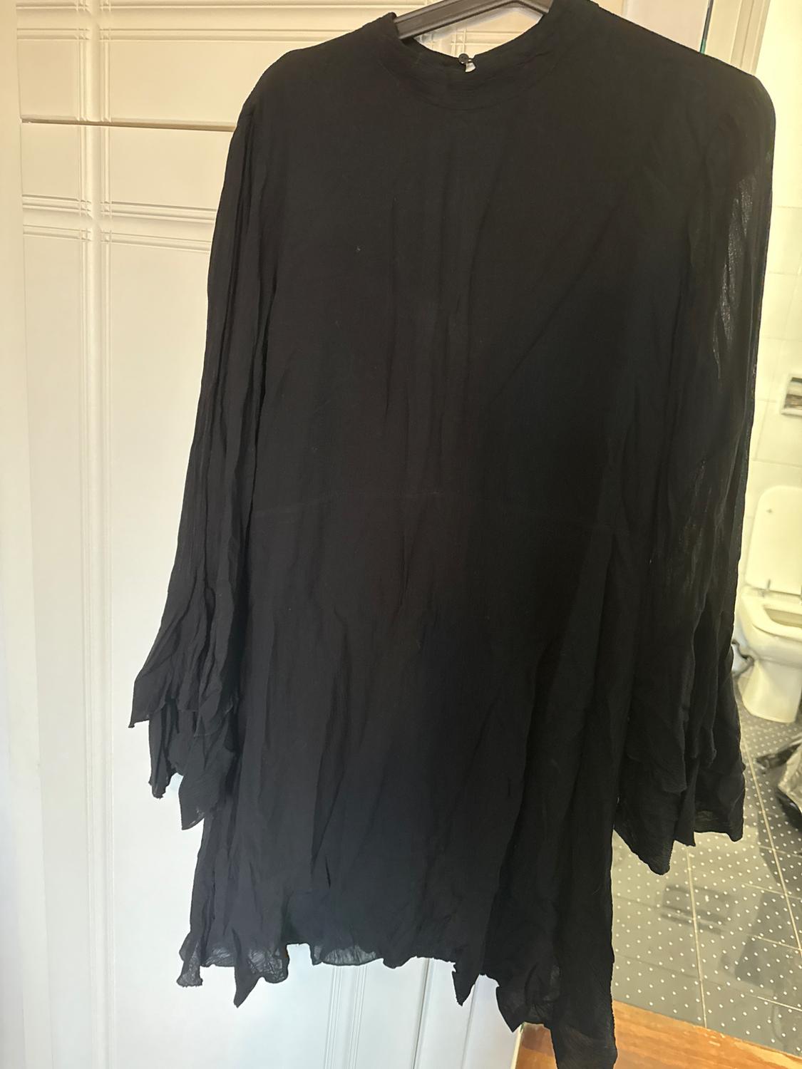 Long-sleeved black dress with a crinkled texture, hanging on a clothes hanger. White wall background with a bathroom visible to the right.
