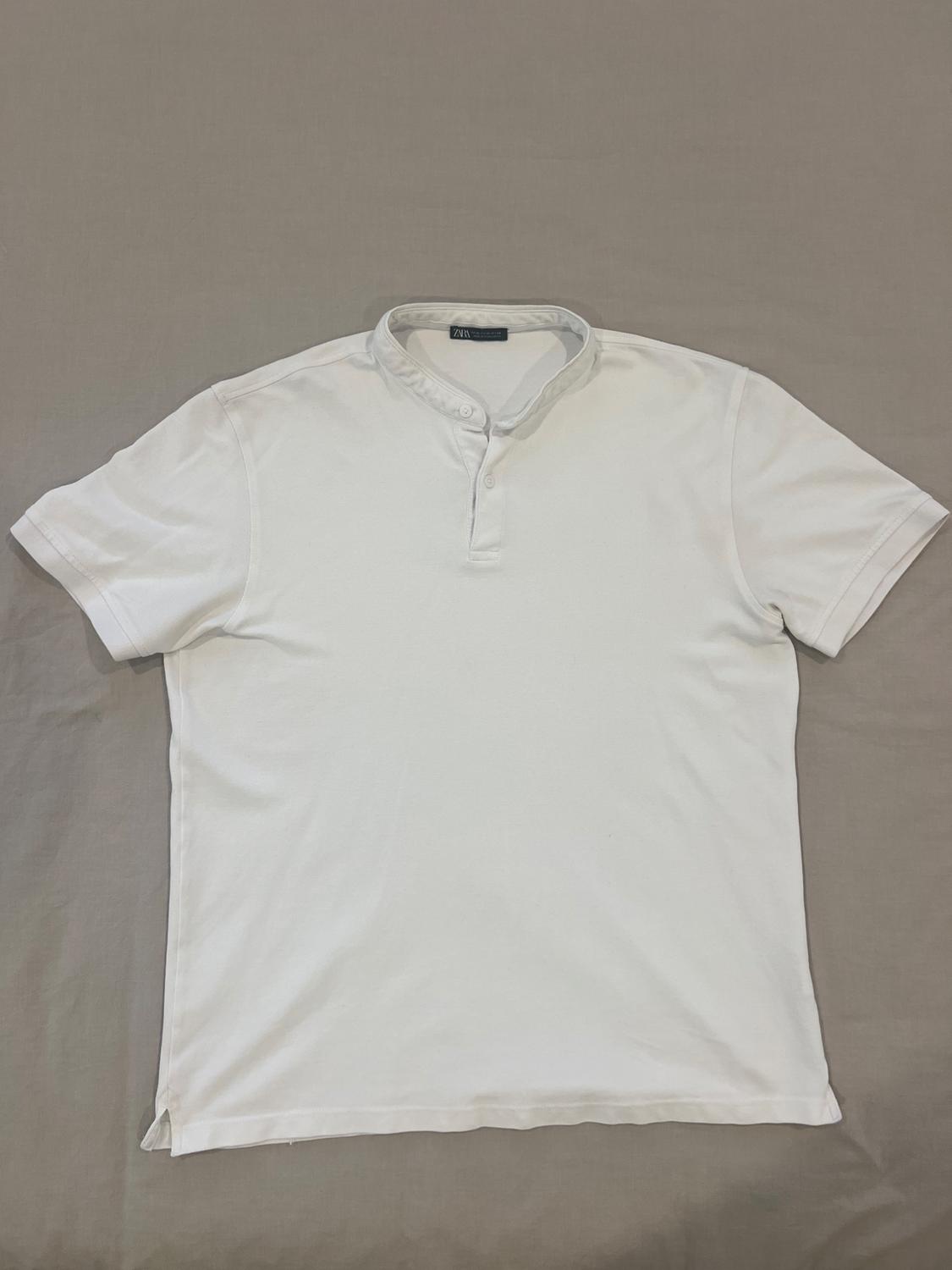 White short-sleeved T-shirt with a grandad collar and three buttons, on a grey background. Visible label on the collar.