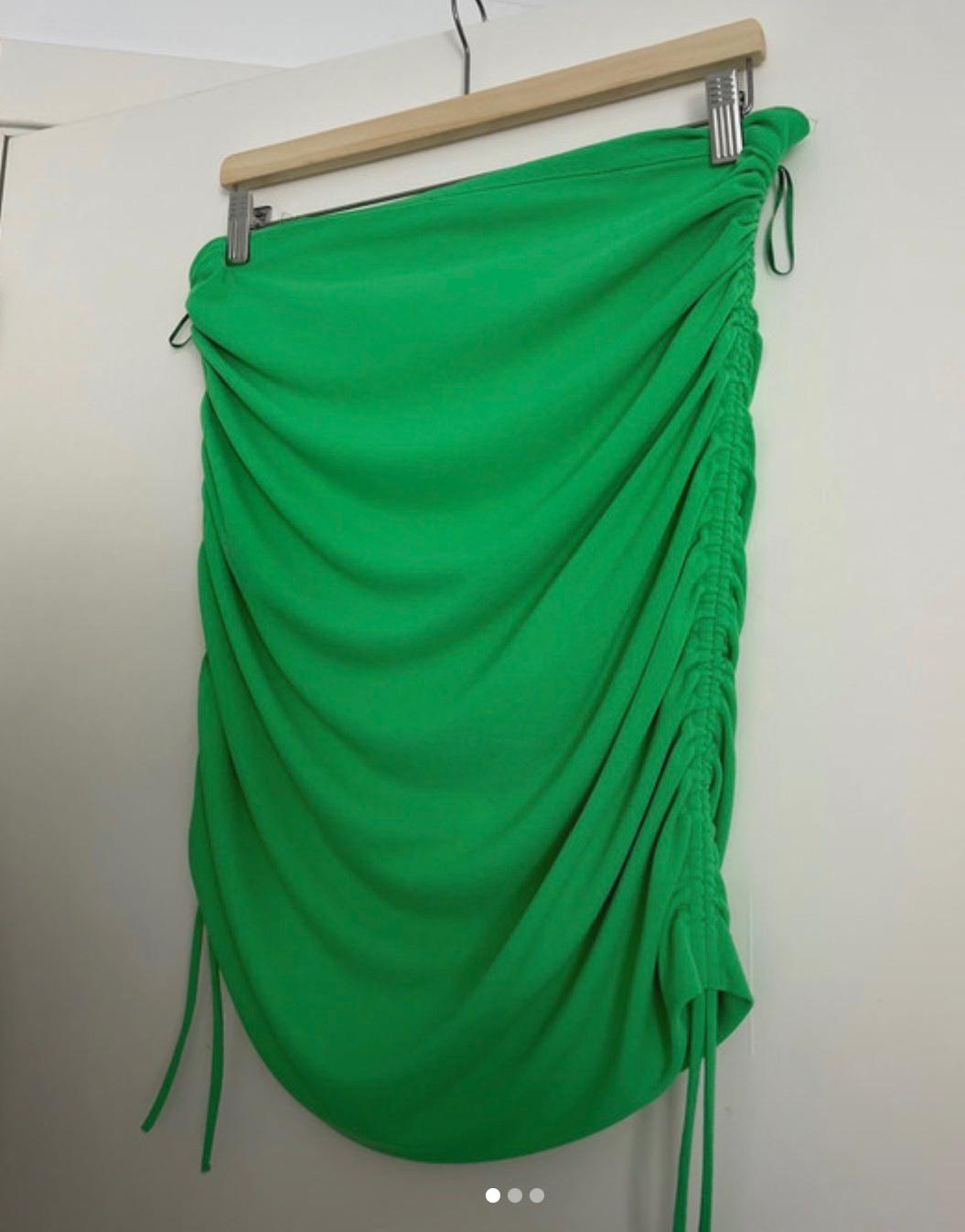 Green draped skirt hanging on a wooden hanger, with ruched details and ties on the sides. White background.