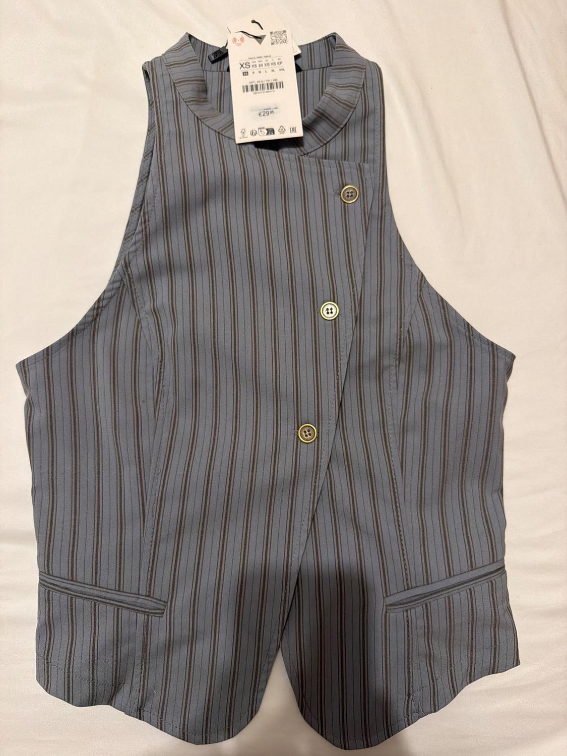 Blue waistcoat with vertical stripes, high neck, asymmetric fastening with three buttons, and pockets. Label with text XS and €29.95.