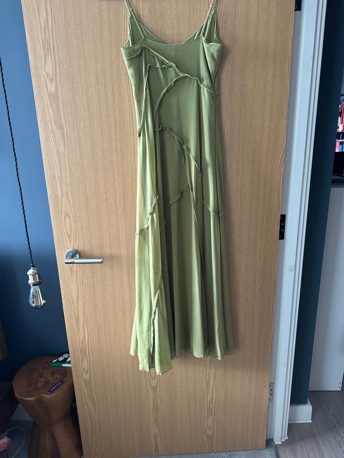 Long green strap dress with asymmetric seams hanging on a wooden door. Blue wall, lamp and the text APERITIVO.