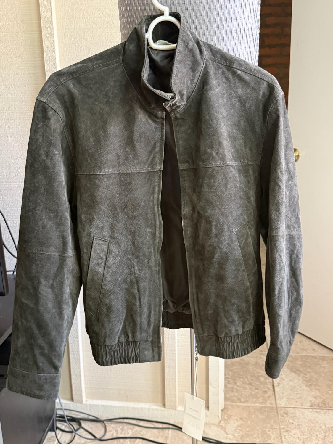 Dark grey jacket hanging on a coat hanger in a domestic interior. It features a stand-up collar, front zip and elasticated trims.