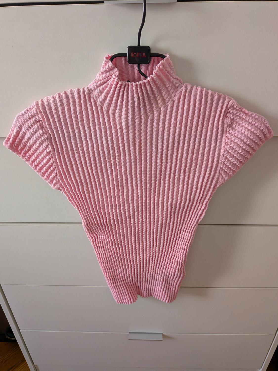 Pink ribbed knit high-neck short-sleeved top, hanging on a black hanger with the text TUMI in front of a white chest of drawers.
