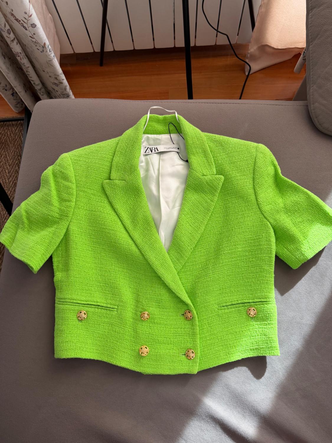 Κοντό blazer σε νέον πράσινο χρώμα με υφή, κοντά μανίκια, χρυσά κουμπιά και ετικέτα ZARA, πάνω σε έναν γκρι καναπέ κάτω από φυσικό φως.