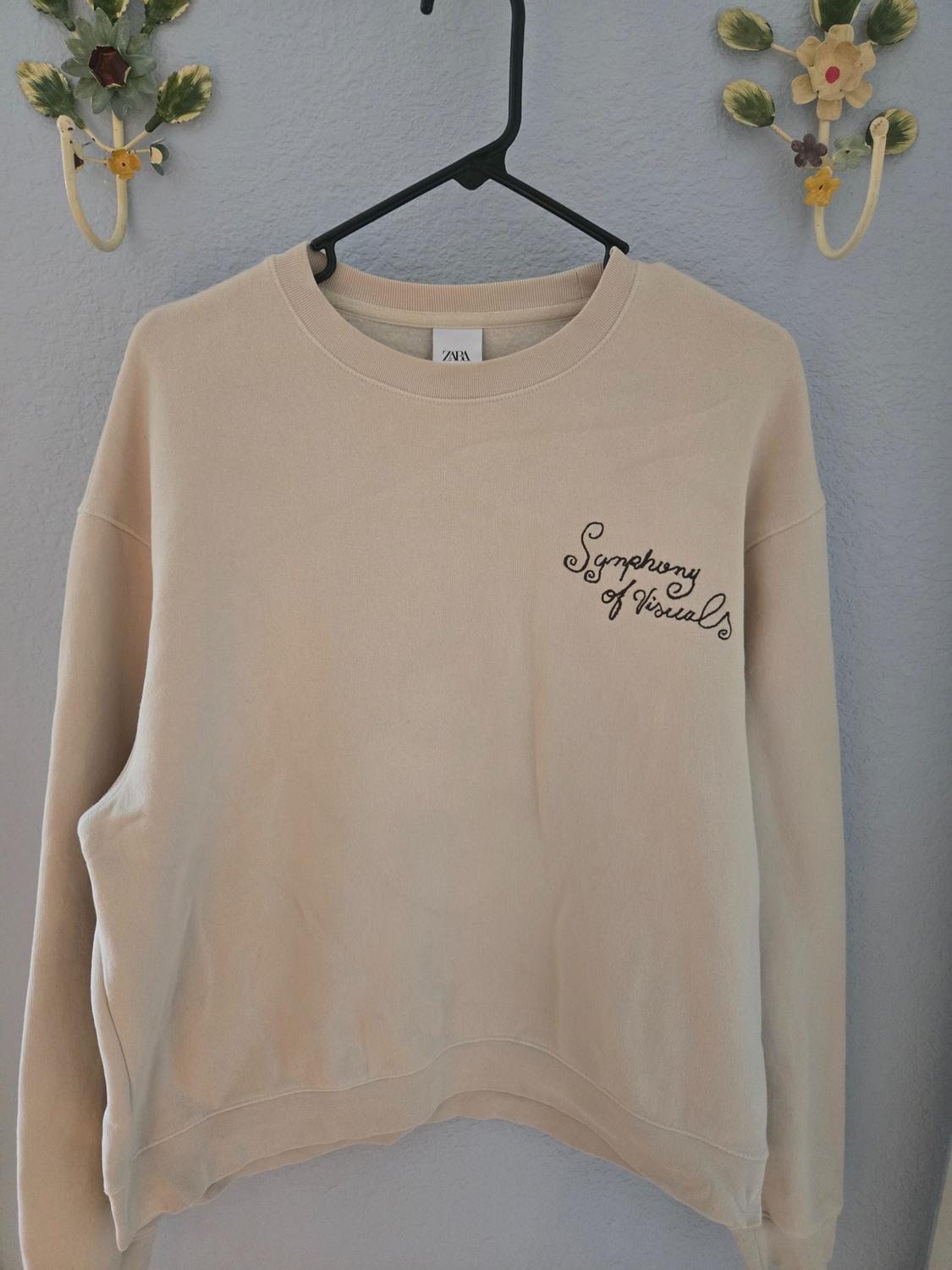 Beige sweatshirt hanging on a black hanger, with the text "Symphony of Visuals" embroidered on the chest. Floral decoration on the wall.