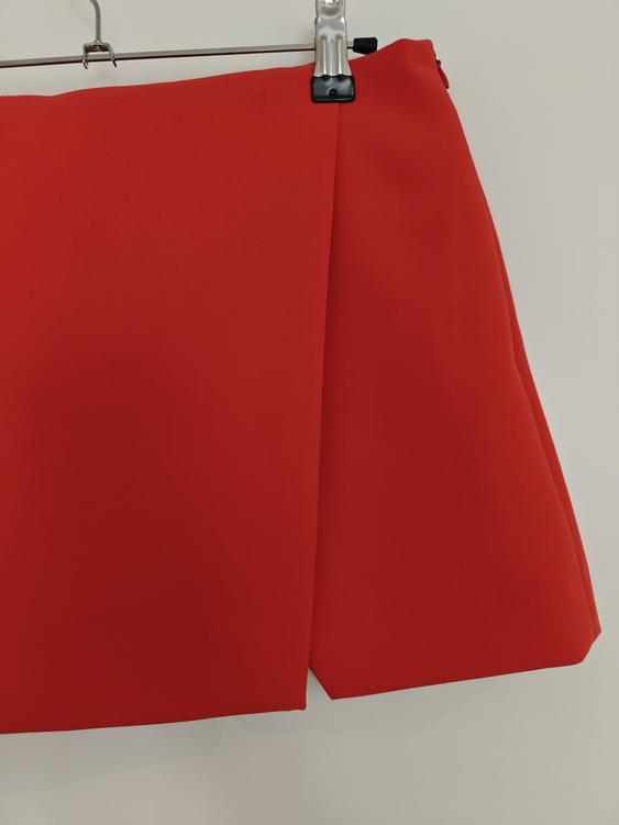 Red skirt hanging on a metal hanger against a white background, showcasing a design with pleats and a straight cut.
