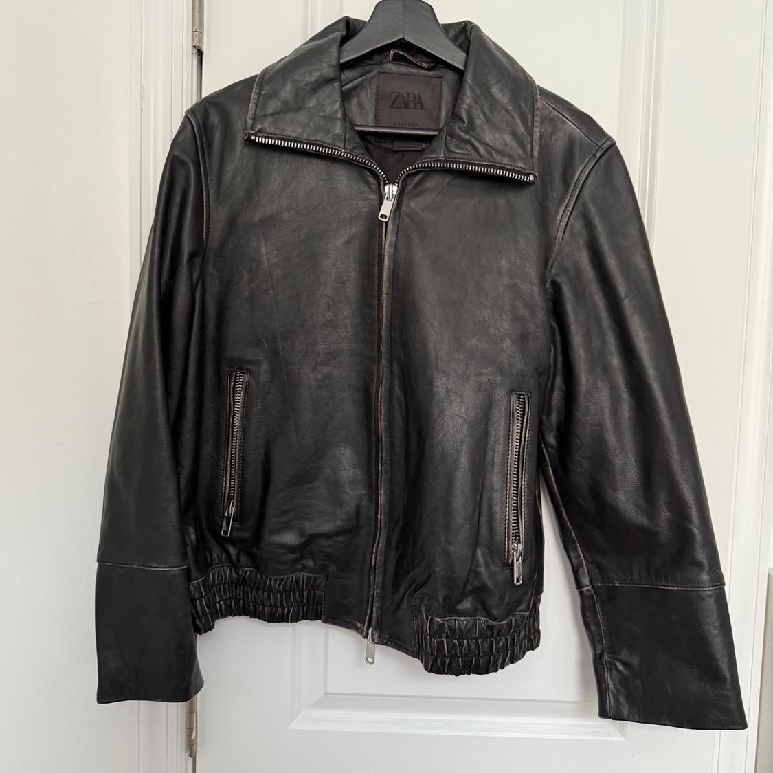 Black leather jacket hanging on a hanger, with a front zipper and side pockets with closures. Visible tag with "ZARA" text.