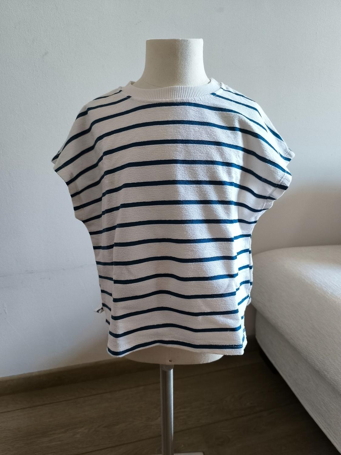 Παιδική κούκλα βιτρίνας με λευκό T-shirt με μπλε οριζόντιες ρίγες. Φωτεινό εσωτερικό με λευκό τοίχο, ξύλινο πάτωμα και λευκό καναπέ.