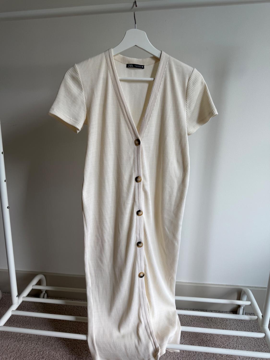 Cream ribbed knit maxi dress with brown buttons down the front, hanging on a white hanger on a metal stand.