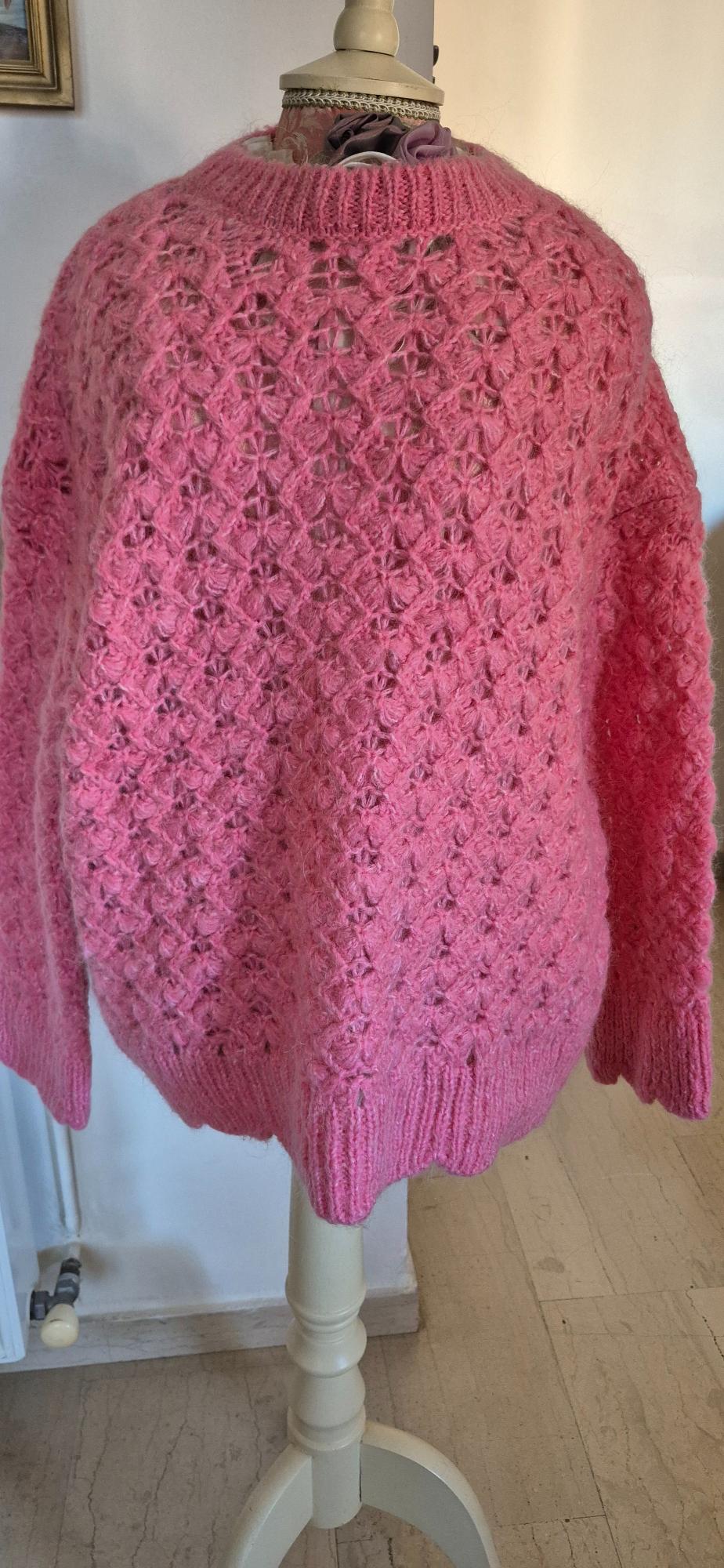 Pink knitted jumper with an openwork design on a mannequin in an indoor setting. Background with a white wall and light marble floor.