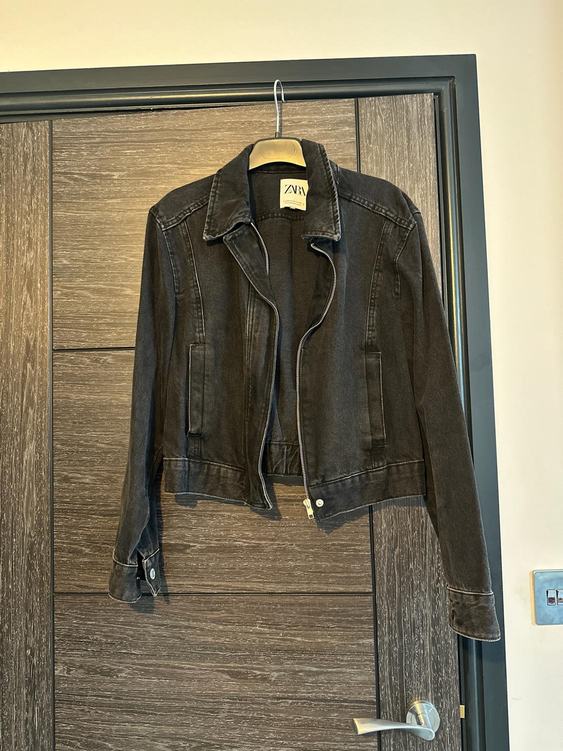 Black denim jacket with a zip hanging on a hanger over a wooden door. Interior label with the text Zara.