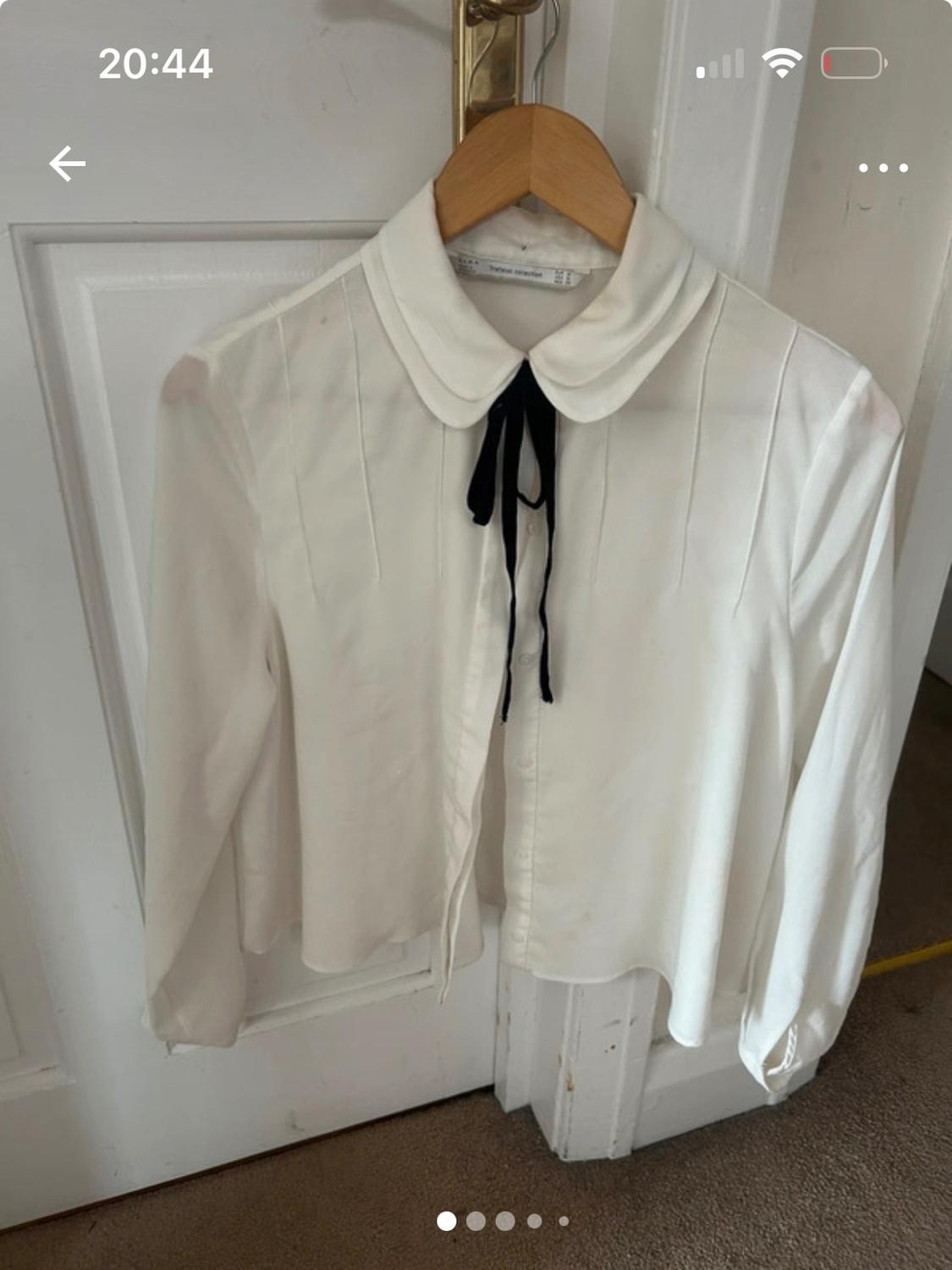White blouse with a round neck and black tie hanging on a wooden hanger over a white door. Mobile phone screen visible.