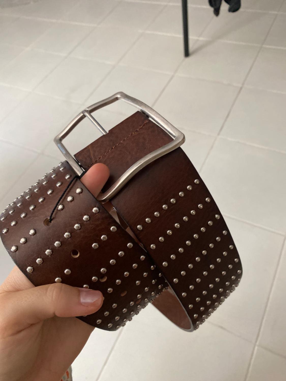A brown belt with silver studs and a metal buckle, held by a hand over a white tiled floor.