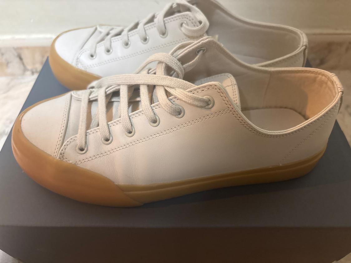 White trainers with a brown rubber sole on a grey box, in an indoor setting with light marble flooring.