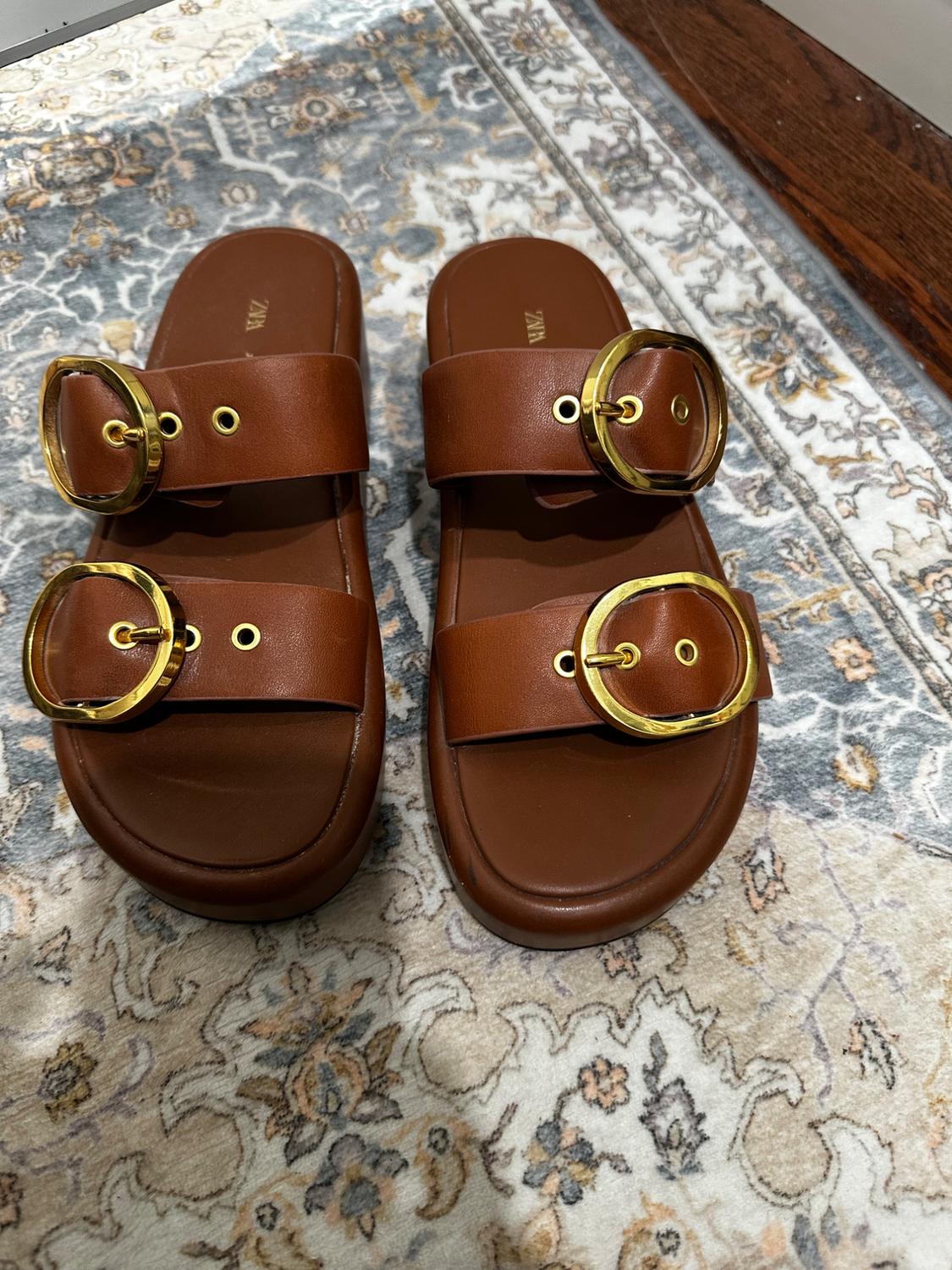 Brown sandals with gold buckles on a floral design rug. Visible text: "ZARA" on the insole.