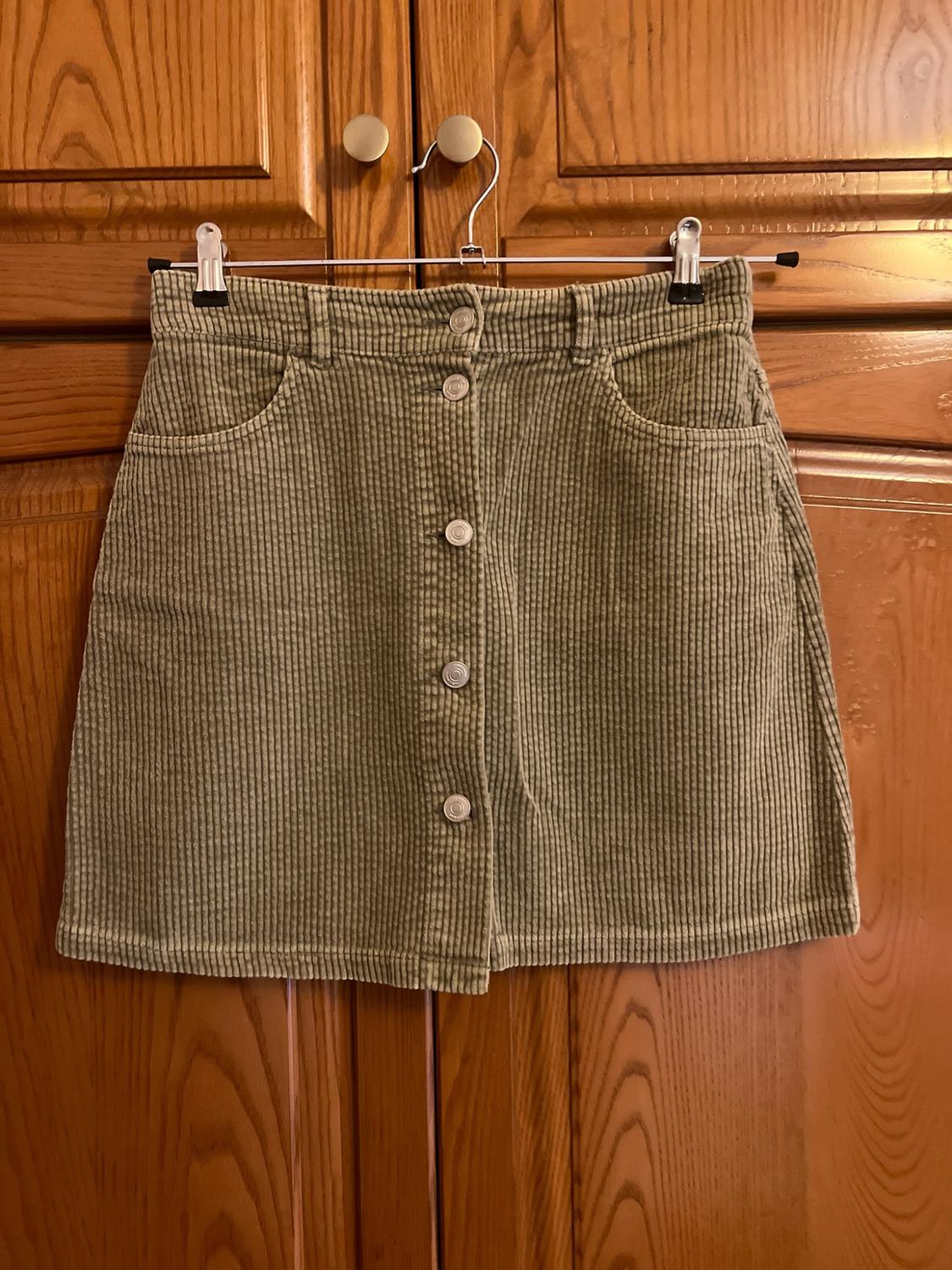 Olive green corduroy mini skirt with front buttons and pockets, hanging on a hanger over a wooden wardrobe. Casual style.