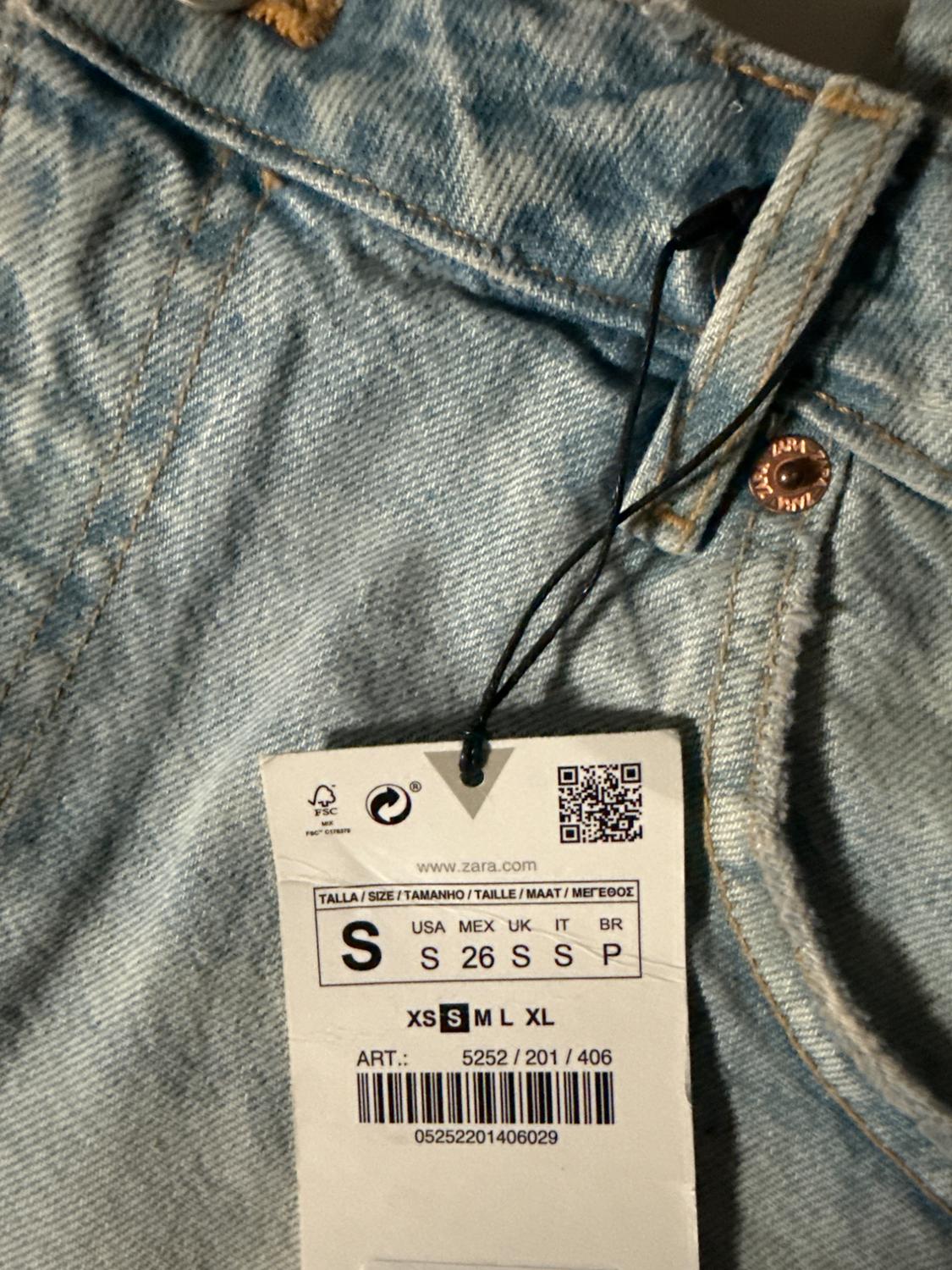 Light blue denim jeans tag with stitching details and a metal button. Visible text: "www.zara.com", size S, barcode.