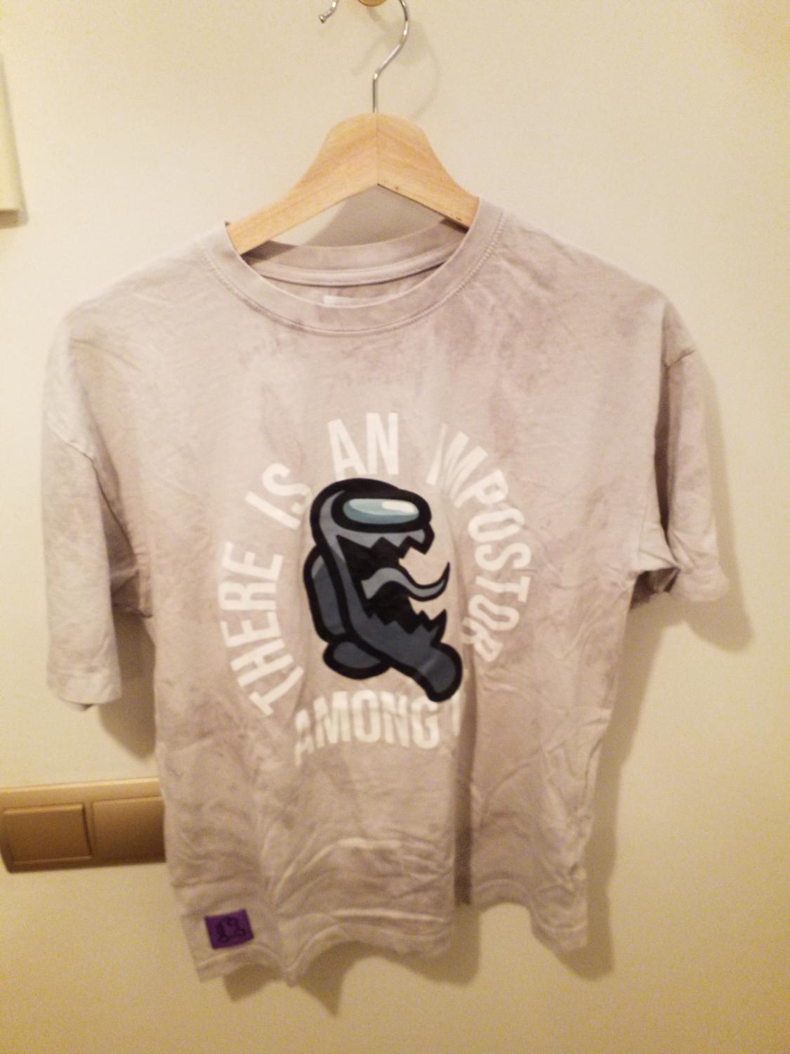 Grey t-shirt hanging on a wooden hanger, with character design and text "THERE IS AN IMPOSTOR AMONG" in white letters.