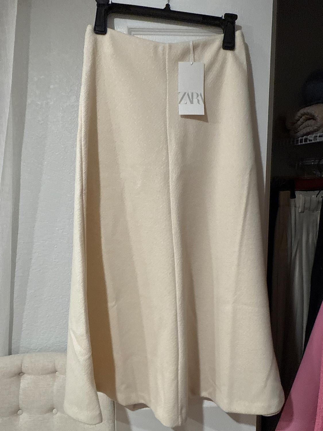 Long beige skirt hanging on a black hanger with a Zara tag, in an indoor setting with a white wall background and a beige chair.