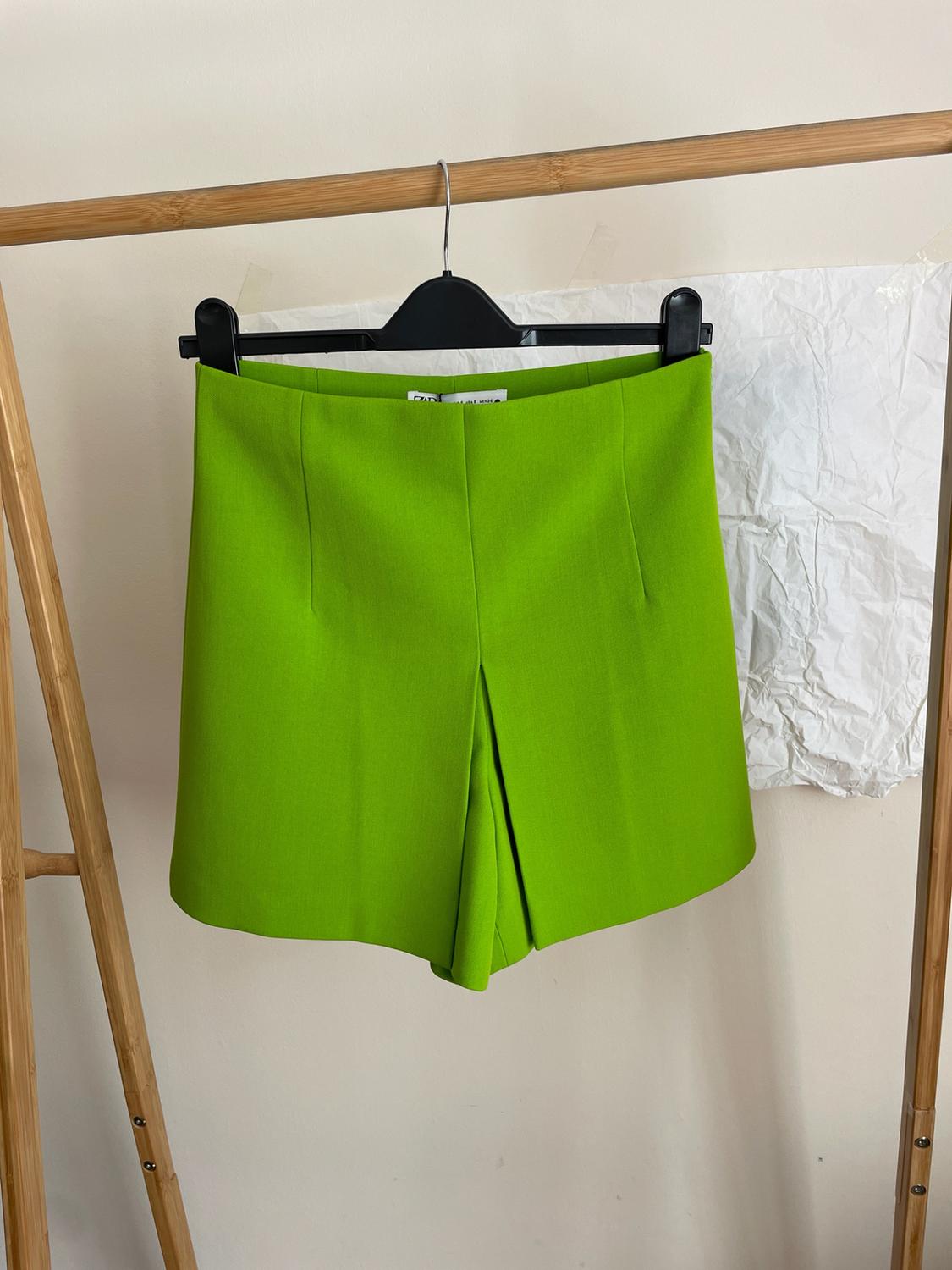 Green skort hanging on a black hanger on a wooden clothes rail. In the background, a light-coloured wall with a crumpled white piece of paper.