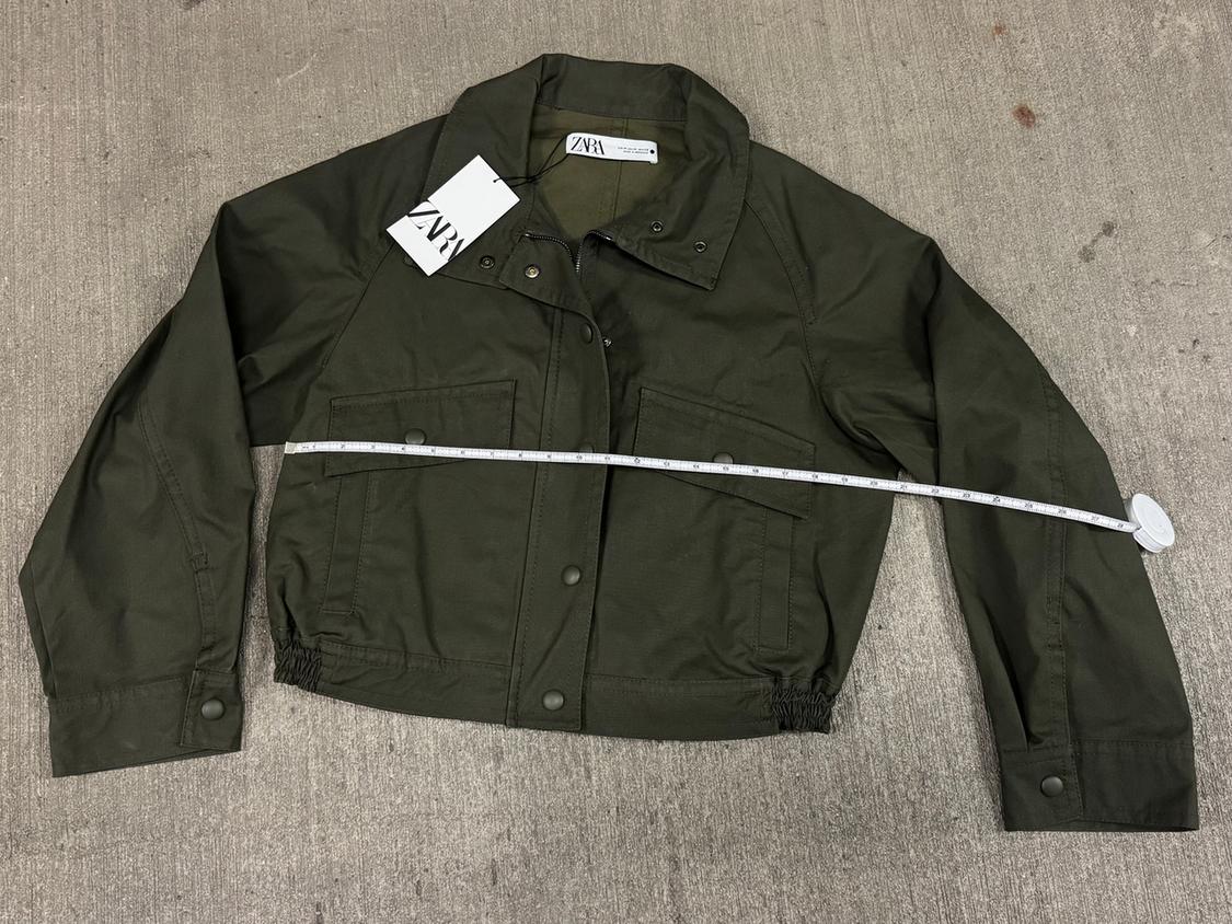 Olive green jacket on a gray background with a white tape measure on top. Visible tag with text "ZARA".