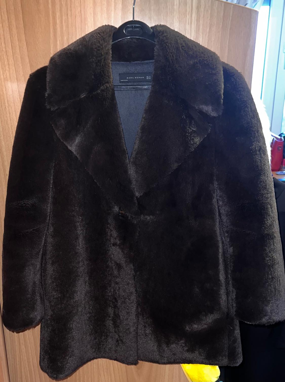 Dark brown faux fur coat hanging on a black hanger, against a light wood background. Label visible inside.