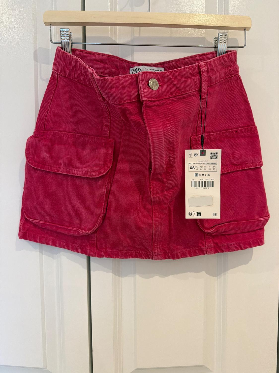Fuchsia pink denim miniskirt with cargo pockets, hanging on a wooden hanger on a white door. Label with the text ZARA.