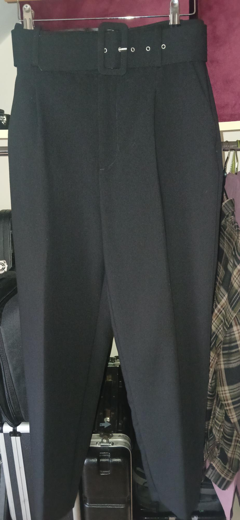 Black trousers hanging on a hanger with a wide belt and square buckle. Background with suitcases and chequered fabric.
