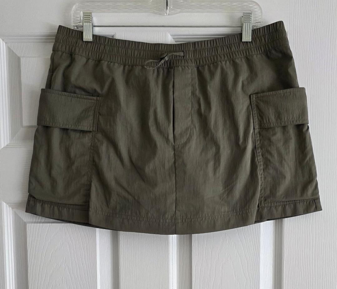Olive green short skirt with large side pockets, hanging on a clear hanger over a white door.