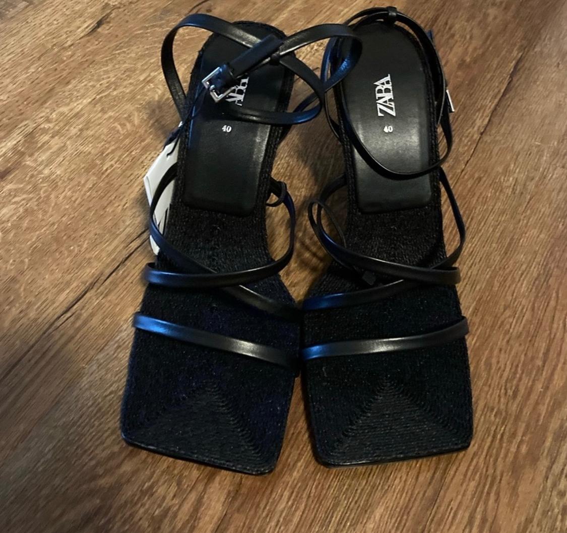 Black square-heeled sandals with thin straps on a wooden floor. Visible text: "ZARA" and "40" on the insole.