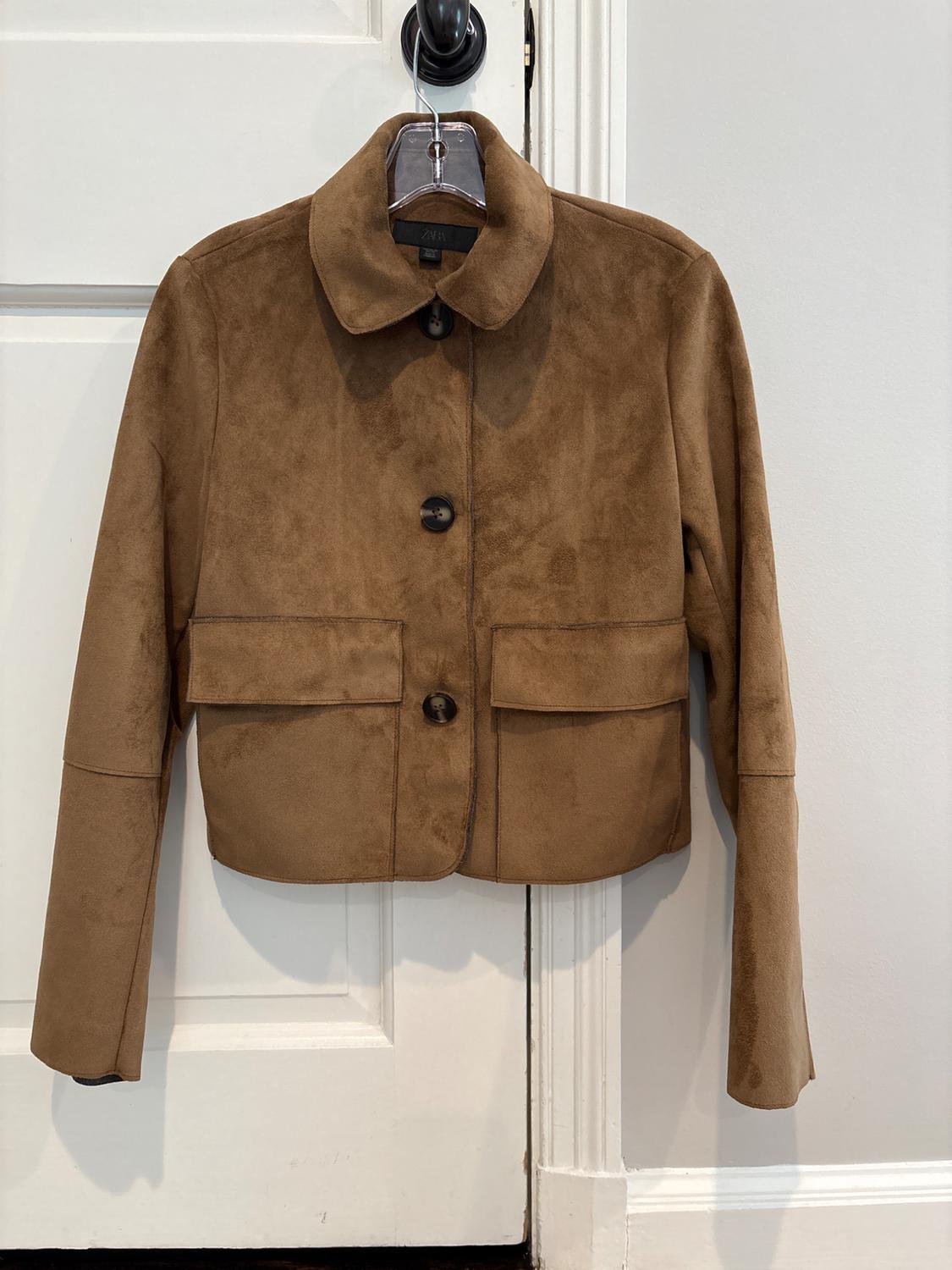 Brown ZARA jacket with a collar, buttons and flap pockets, hanging on a clear hanger over a white door.