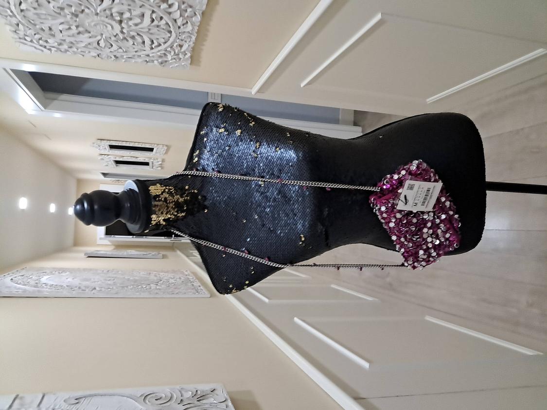 Black mannequin with gold sequins, small purple bag with chain and tag, in a hallway with white embossed walls.