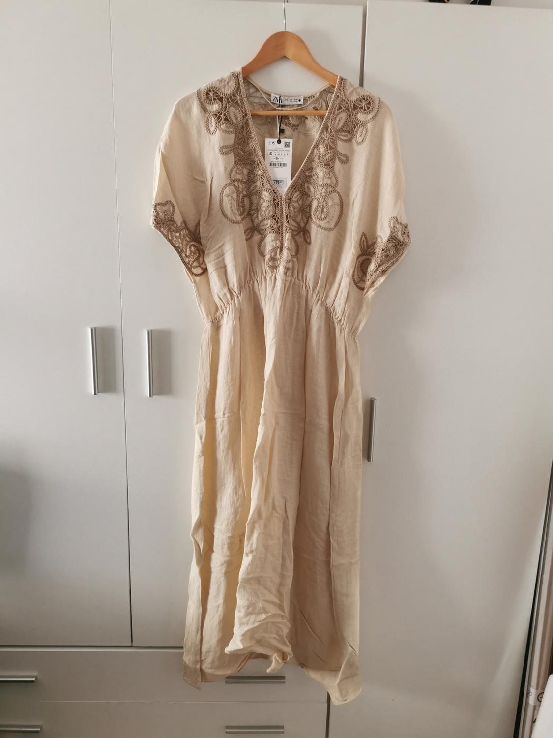 Beige relaxed-fit dress, V-neck, ruched waist, and brown embroidery. Hanging in white wardrobe. Text: ZARA, S.
